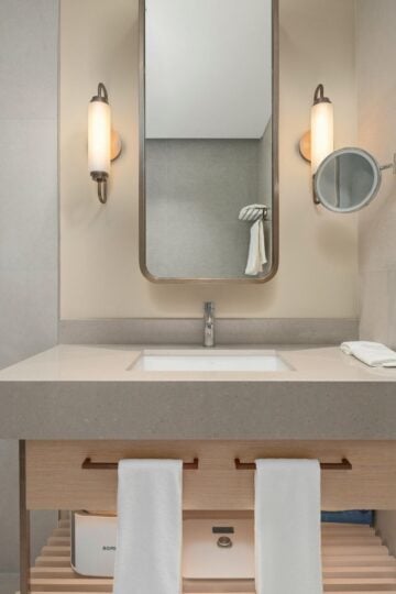 Modern bathroom vanity with a rectangular basin, wall-mounted mirror, two wall lights, a magnifying mirror, and two white towels hanging on a wooden cupboard.