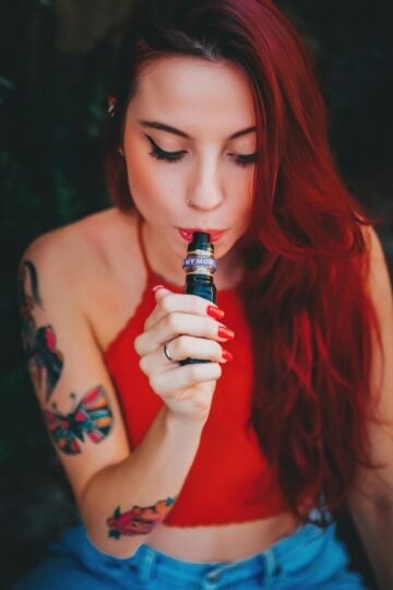 A woman with long red hair and tattoos sits outdoors, holding a 10 Motives Vape to her mouth, enjoying the simplicity and satisfaction it brings, whilst wearing a red sleeveless top and blue jeans.