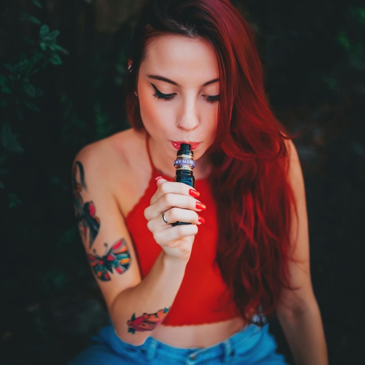 Why 10 Motives Vape Is a Go-To Choice for Simplicity and Satisfaction A woman with long red hair and tattoos sits outdoors, holding a 10 Motives Vape to her mouth, enjoying the simplicity and satisfaction it brings, whilst wearing a red sleeveless top and blue jeans.