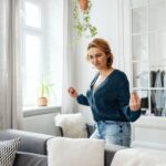 How Light Affects Your Mood (and What to Do About It) A woman with short hair wearing a blue jumper and jeans stands in a bright living room, making a playful gesture with her hands.