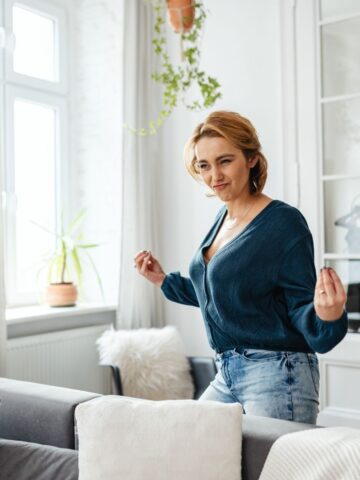 A woman with short hair wearing a blue jumper and jeans stands in a bright living room, making a playful gesture with her hands.