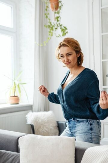 A woman with short hair wearing a blue jumper and jeans stands in a bright living room, making a playful gesture with her hands.