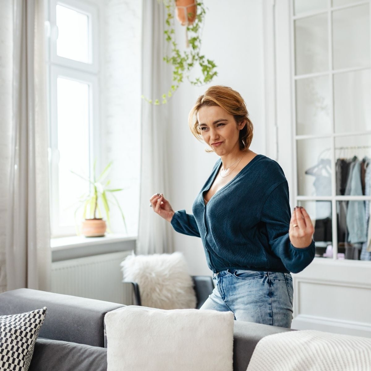 How Light Affects Your Mood (and What to Do About It) A woman with short hair wearing a blue jumper and jeans stands in a bright living room, making a playful gesture with her hands.