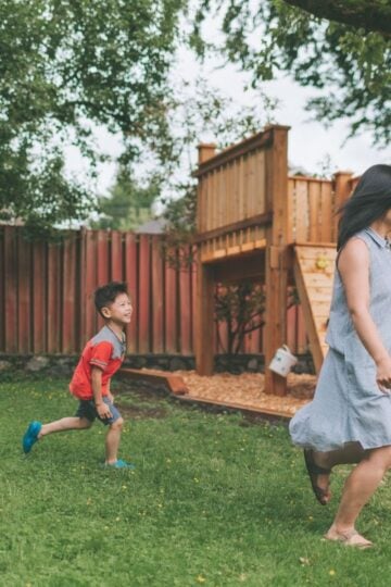 How Families Are Redesigning Their Gardens for Year-Round Play A woman in a light dress runs on grass whilst a young boy in a red shirt follows her near a wooden play structure in a back garden, enjoying year-round play amid the greenery of family gardens.