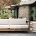 Outdoor patio with a light-coloured cushioned sofa, a round side table, and plants creates a welcoming outdoor living space in front of a modern stone house.