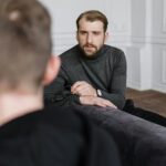 The Growing Importance of Accessible Behavioral Health Services A man with a beard in a grey polo neck sits on a sofa, holding spectacles and looking at another person whose back is to the camera, illustrating the importance of accessible behavioural health services in a supportive setting.