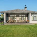 Easy Ways to Keep Your Yard Fresh and Green This Summer Single-storey modern house with large windows, a covered patio featuring outdoor furniture, and a well-maintained green lawn-perfect for showcasing your garden care skills and trying out summer lawn tips under a clear blue sky.