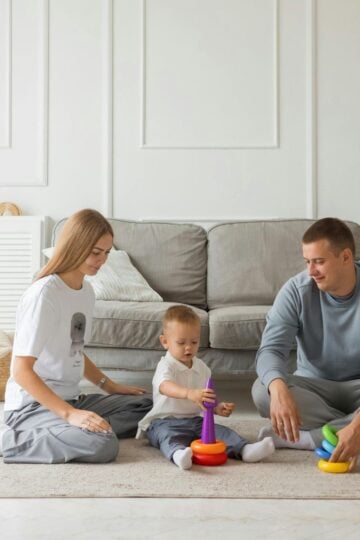 A woman, a child, and a man sit on the floor in a busy family home, playing with colourful stacking rings on a beige rug in front of a grey sofa.