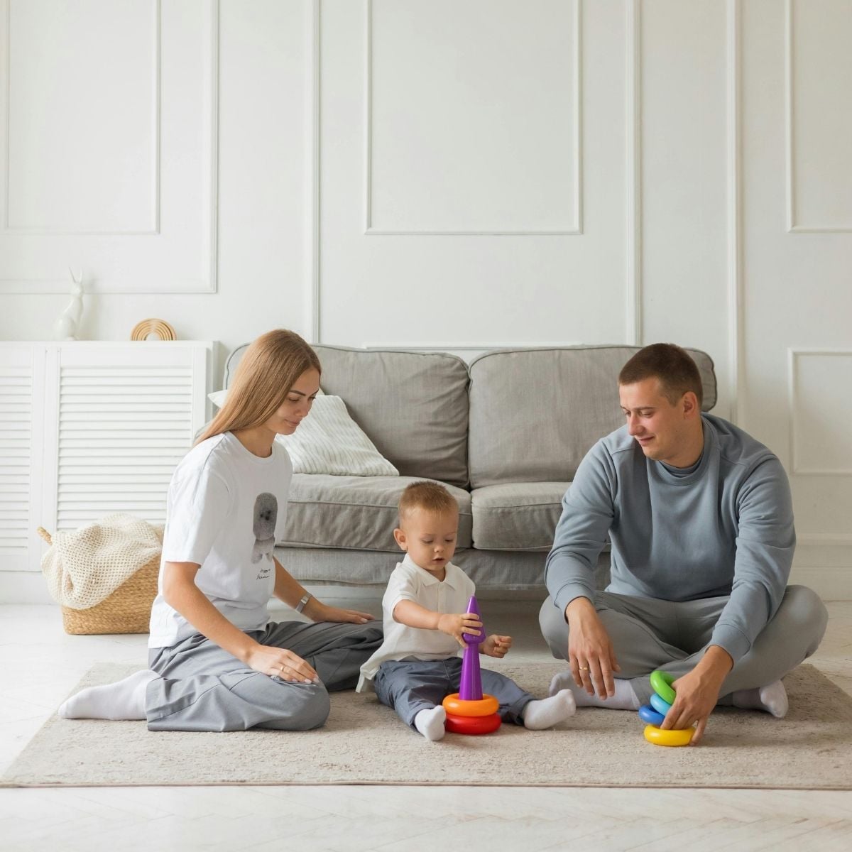 Balanced Routines: Practical Ways to Keep a Busy Family Home Calm and Restful A woman, a child, and a man sit on the floor in a busy family home, playing with colourful stacking rings on a beige rug in front of a grey sofa.