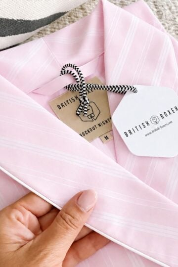 A hand holds a folded pink and white striped pyjama top with tags reading "British Boxers" and "Luscout Nightshirt M," perfect for pairing with a Satin Stripe Dressing Gown from British Boxers.