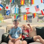 How To Host A Football Watch Party At Home This Summer Two boys sit on a sofa in football kits, hosting a football watch party at home, with colourful strings of international flags hanging behind them.