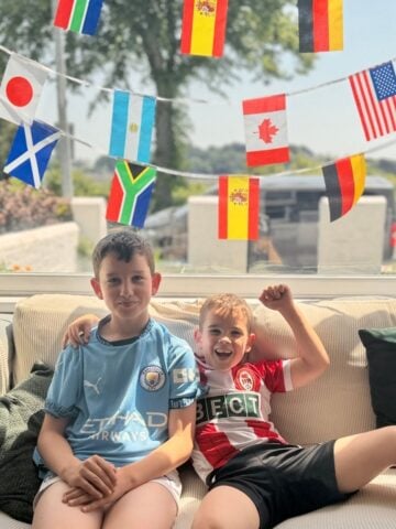 How To Host A Football Watch Party At Home This Summer Two boys sit on a sofa in football kits, hosting a football watch party at home, with colourful strings of international flags hanging behind them.