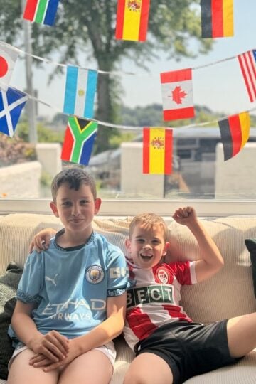 Two boys sit on a sofa in football kits, hosting a football watch party at home, with colourful strings of international flags hanging behind them.