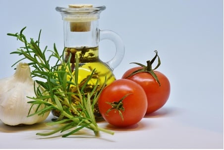 Can You Cook With Olive Oil? A glass bottle of cooking olive oil, two tomatoes, a bulb of garlic, and sprigs of rosemary are arranged on a white surface.