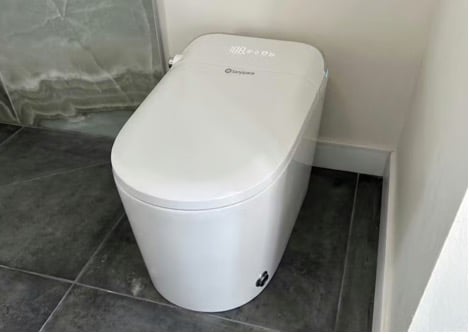 A modern, white smart toilet with digital controls is installed in a bathroom with dark tiled flooring and light-coloured walls.