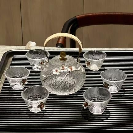 A glass teapot with a decorated handle and floral design is placed on a tray, surrounded by six matching glass teacups.