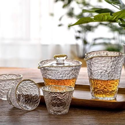 A textured glass jug and matching cups with amber liquid are arranged on a wooden tray, with green plants in the background.