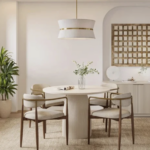 A modern dining space featuring a round white dining table, four upholstered chairs, a pendant light, potted plant, and decorative items on a sideboard against a beige wall.