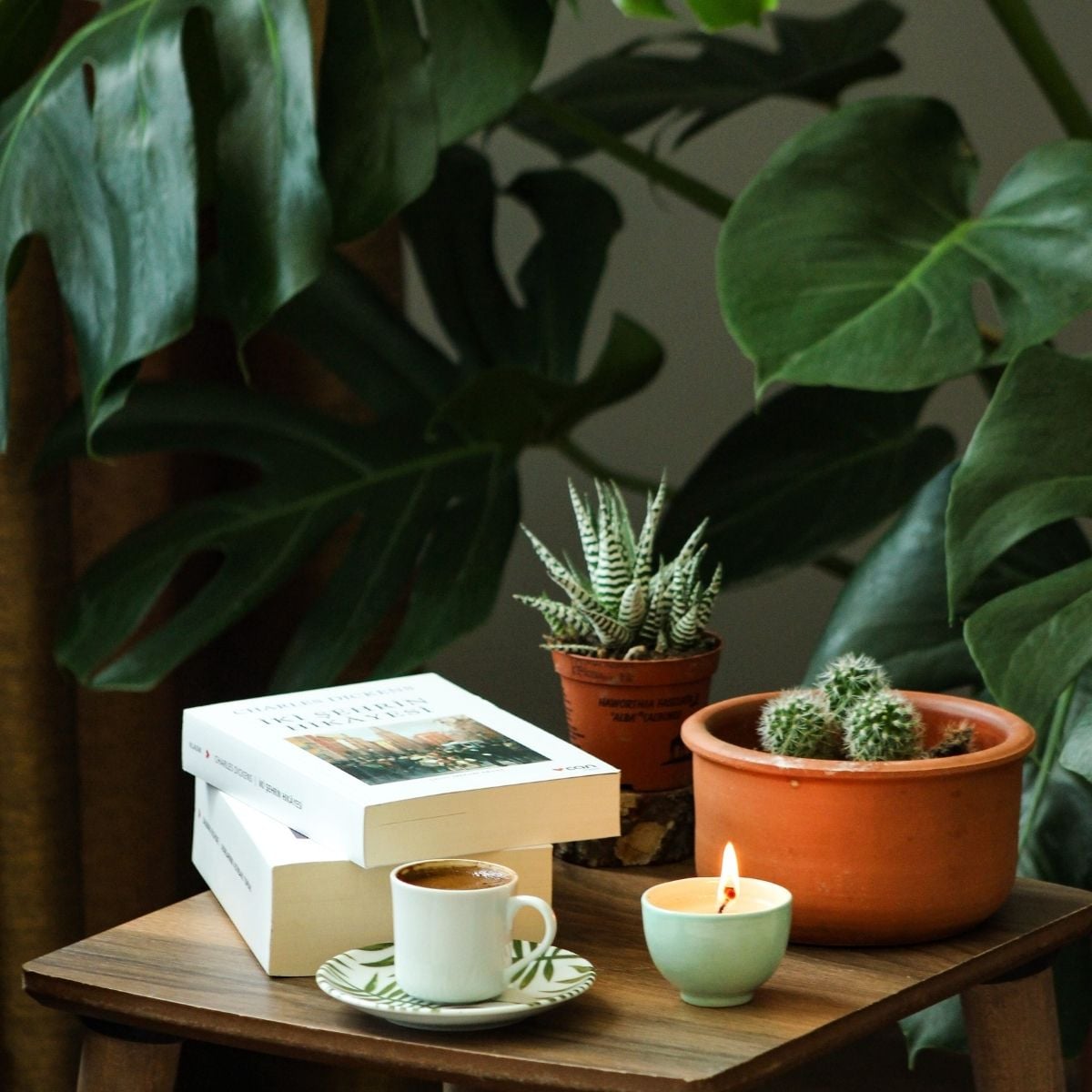 How to Create a Calmer Home: Simple Daily Rituals That Support Wellbeing A wooden table holds two books, a cup of coffee on a saucer, a lit candle, and two potted succulents—inviting daily rituals that foster wellbeing amid large green leaves in the background.