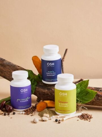 Three OSH Wellness supplement bottles are arranged on a log with various natural ingredients and leaves on a beige background, reflecting the care taken to choose quality supplements for your family's wellbeing.