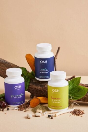 Three OSH Wellness supplement bottles are arranged on a log with various natural ingredients and leaves on a beige background, reflecting the care taken to choose quality supplements for your family’s wellbeing.