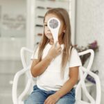 A young girl with long hair sits on a white chair, holding an occluder over one eye during a comfortable eye care examination by a home visit optician.