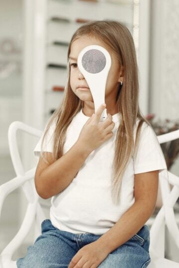 A young girl with long hair sits on a white chair, holding an occluder over one eye during a comfortable eye care examination by a home visit optician.