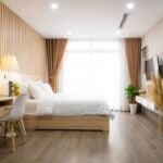 Modern bedroom with a large bed, white bedding, wooden floor, desk and chair, potted plants, TV, and floor-to-ceiling window with brown curtains-ideal for letting in Georgia while enjoying natural light and comfort.