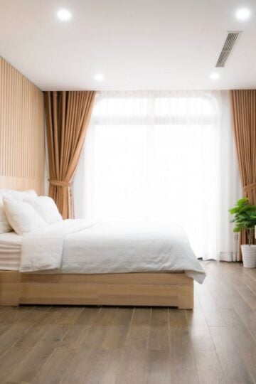 A Tenant’s Guide to Renting in Georgia: Know Your Rights and Responsibilities Modern bedroom with a large bed, white bedding, wooden floor, desk and chair, potted plants, TV, and floor-to-ceiling window with brown curtains—ideal for letting in Georgia while enjoying natural light and comfort.