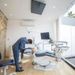 The Process Behind a Complete Dental Treatment Plan and What Patients Should Expect A dentist in a suit leans over a patient reclined in a dental chair inside a modern, well-lit dental surgery with large windows, discussing the dental treatment plan to ensure patient expectations are met.