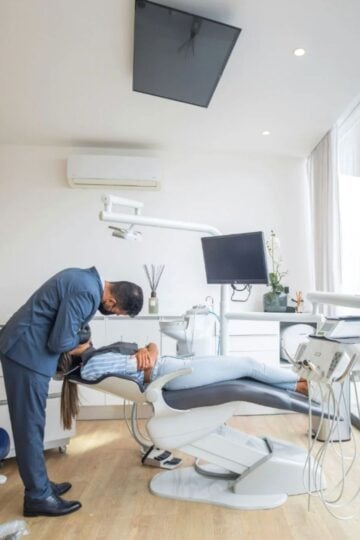 The Process Behind a Complete Dental Treatment Plan and What Patients Should Expect A dentist in a suit leans over a patient reclined in a dental chair inside a modern, well-lit dental surgery with large windows, discussing the dental treatment plan to ensure patient expectations are met.