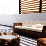 How to Create the Perfect Outdoor Living Space for Families Create a cosy outdoor living space for families with a wicker sofa featuring white cushions and a matching round glass-topped table, set on a patio with a wooden slat privacy screen and white walls.