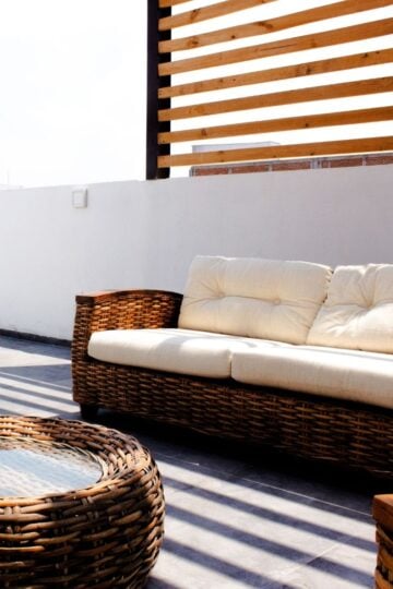 How to Create the Perfect Outdoor Living Space for Families Create a cosy outdoor living space for families with a wicker sofa featuring white cushions and a matching round glass-topped table, set on a patio with a wooden slat privacy screen and white walls.