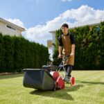 Step-by-Step Guide to Replacing Lawn Mower Blades A person wearing an apron is pushing a lawnmower across a grassy garden on a sunny day, with hedges and houses in the background-perfect for those following a step-by-step guide to replacing lawnmower blades.
