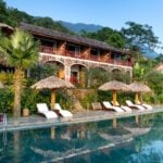 A tropical resort offering affordable luxury, with a two-storey rustic building, lounge chairs, and thatched umbrellas beside a pool, all surrounded by lush greenery and mountains in the background.