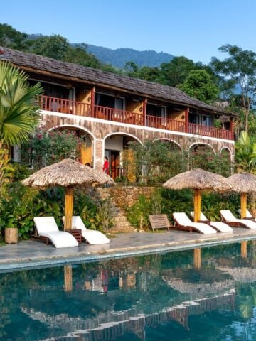 A tropical resort offering affordable luxury, with a two-storey rustic building, lounge chairs, and thatched umbrellas beside a pool, all surrounded by lush greenery and mountains in the background.