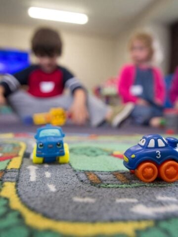 Two toy cars rest on a road-themed play mat in an Early Childhood Environments setting, whilst three children play together in the blurred background at River Oaks.