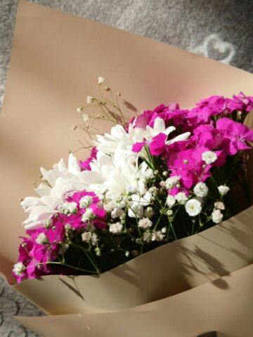 Top Flower Delivery Services in Russia for International Customers A bouquet of white daisies, pink flowers, and baby's breath wrapped in beige paper, placed on a patterned grey surface in sunlight. Perfect for sending love with Flower Delivery Russia.