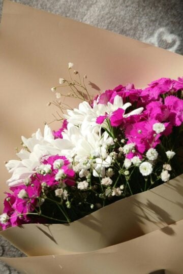 A bouquet of white daisies, pink flowers, and baby's breath wrapped in beige paper, placed on a patterned grey surface in sunlight. Perfect for sending love with Flower Delivery Russia.