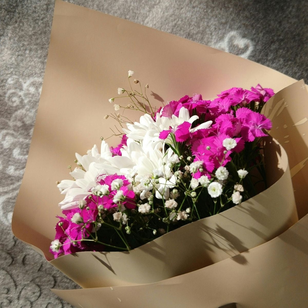 Top Flower Delivery Services in Russia for International Customers A bouquet of white daisies, pink flowers, and baby's breath wrapped in beige paper, placed on a patterned grey surface in sunlight. Perfect for sending love with Flower Delivery Russia.