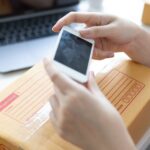 What to Do With Your Old Phone (Before It Ends Up Forgotten in a Drawer) Person holding an old phone above a sealed cardboard parcel with shipping label, next to a laptop-showing one way to recycle old mobile devices responsibly.