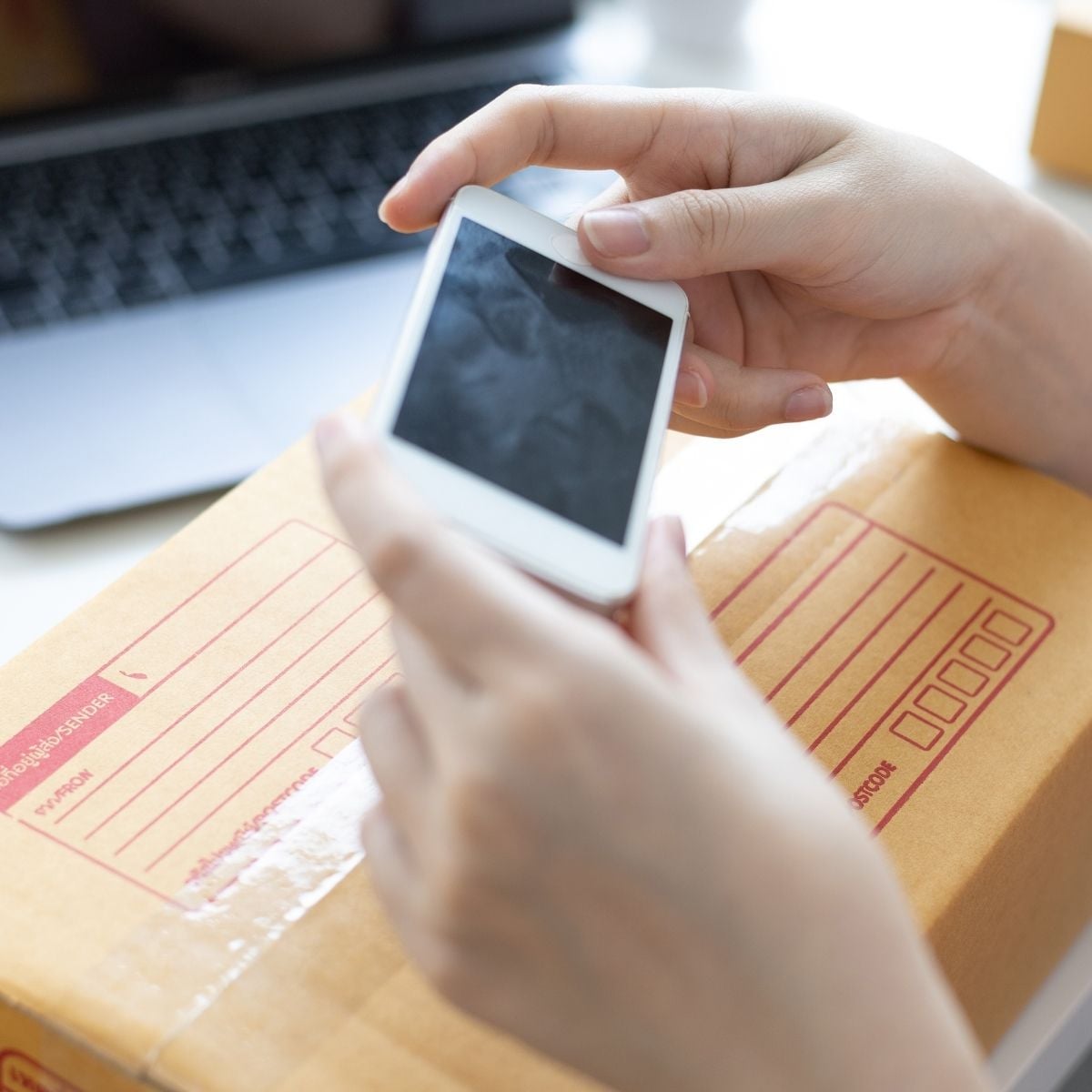 What to Do With Your Old Phone (Before It Ends Up Forgotten in a Drawer) Person holding an old phone above a sealed cardboard parcel with shipping label, next to a laptop—showing one way to recycle old mobile devices responsibly.