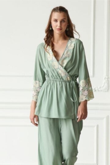 Woman embracing Ordinary Moments in a sage green wrap top with lace detail on the sleeves and matching wide-leg trousers, standing against a white ornate wall—perfect loungewear for serene days at home.
