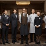 From Five-Star Luxury to Casual Retreats: How to Choose Hotel Uniforms That Reflect Your Brand A group of five hotel staff members in luxury hotel uniforms stand in a line in an elegant, wood-panelled room with floral arrangements in the background, reflecting a strong brand identity.