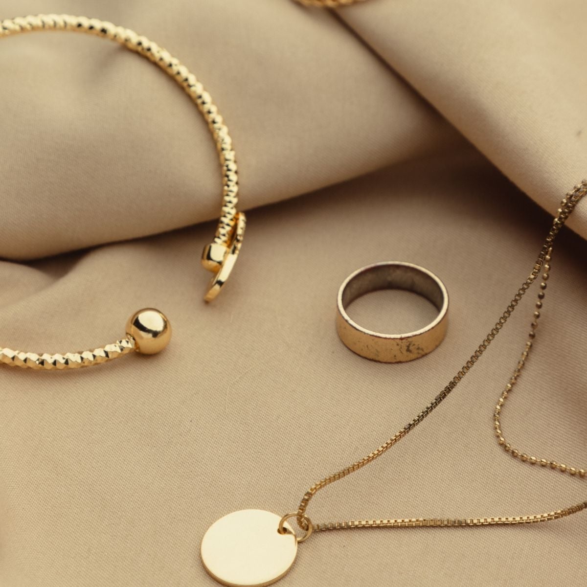 The Jewellery Packing Guide: What to Take (And What to Leave at Home) A gold bracelet, a plain gold ring, and a gold necklace with a round pendant are arranged on beige fabric—a perfect display for jewellery packing inspiration or handy jewellery travel tips.