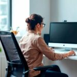 What to Look for When Choosing an Ergonomics Company A woman wearing glasses sits at a desk working on a computer in a bright, modern office, benefiting from ergonomics services provided by a leading ergonomics company.