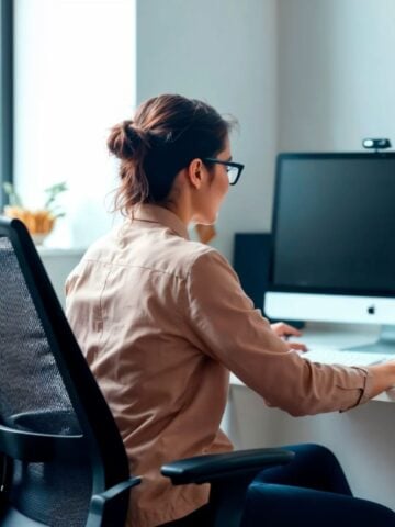 What to Look for When Choosing an Ergonomics Company A woman wearing glasses sits at a desk working on a computer in a bright, modern office, benefiting from ergonomics services provided by a leading ergonomics company.