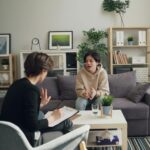 Two people are having a conversation in a living room; one, possibly a parent, is taking notes on a clipboard while the other speaks from the sofa-perhaps discussing trauma treatment for teenagers and ways to find support.