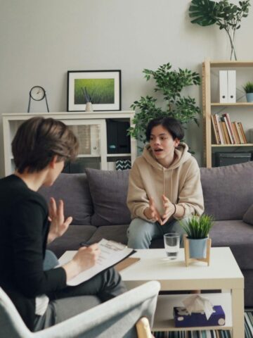Two people are having a conversation in a living room; one, possibly a parent, is taking notes on a clipboard while the other speaks from the sofa-perhaps discussing trauma treatment for teenagers and ways to find support.