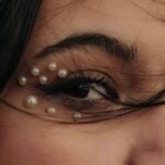 The 5-Minute Eyebrow Routine That Changes Your Whole Face A close-up of a person's eye with small decorative pearls around the eyelid and lower eye area, strands of hair partially covering the face-a stunning facial transformation perfect for your next five-minute beauty routine.